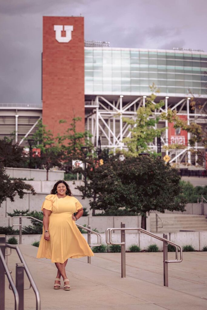 Bethany Hardwig, a curvy black woman wearing a yellow dress, stands on the steps opposite the University of Utah football stadium